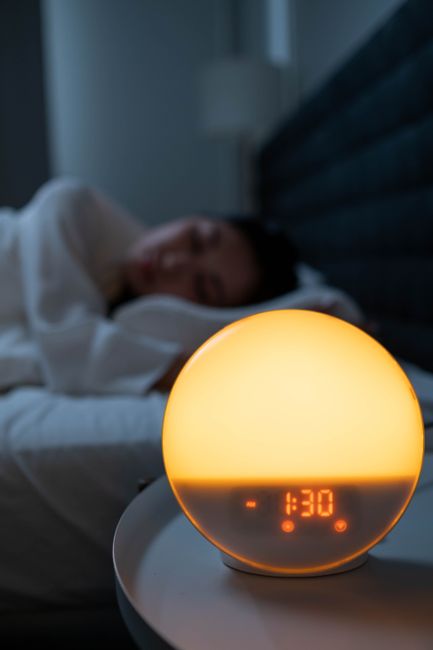 A round sunrise alarm lamp glowing orange on a bedside table beside a woman asleep in bed