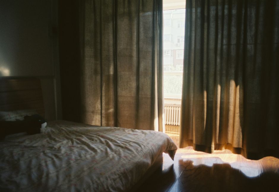 Daylight streaming through partly drawn curtains into a dark bedroom, with a patch of sunlight on the floor